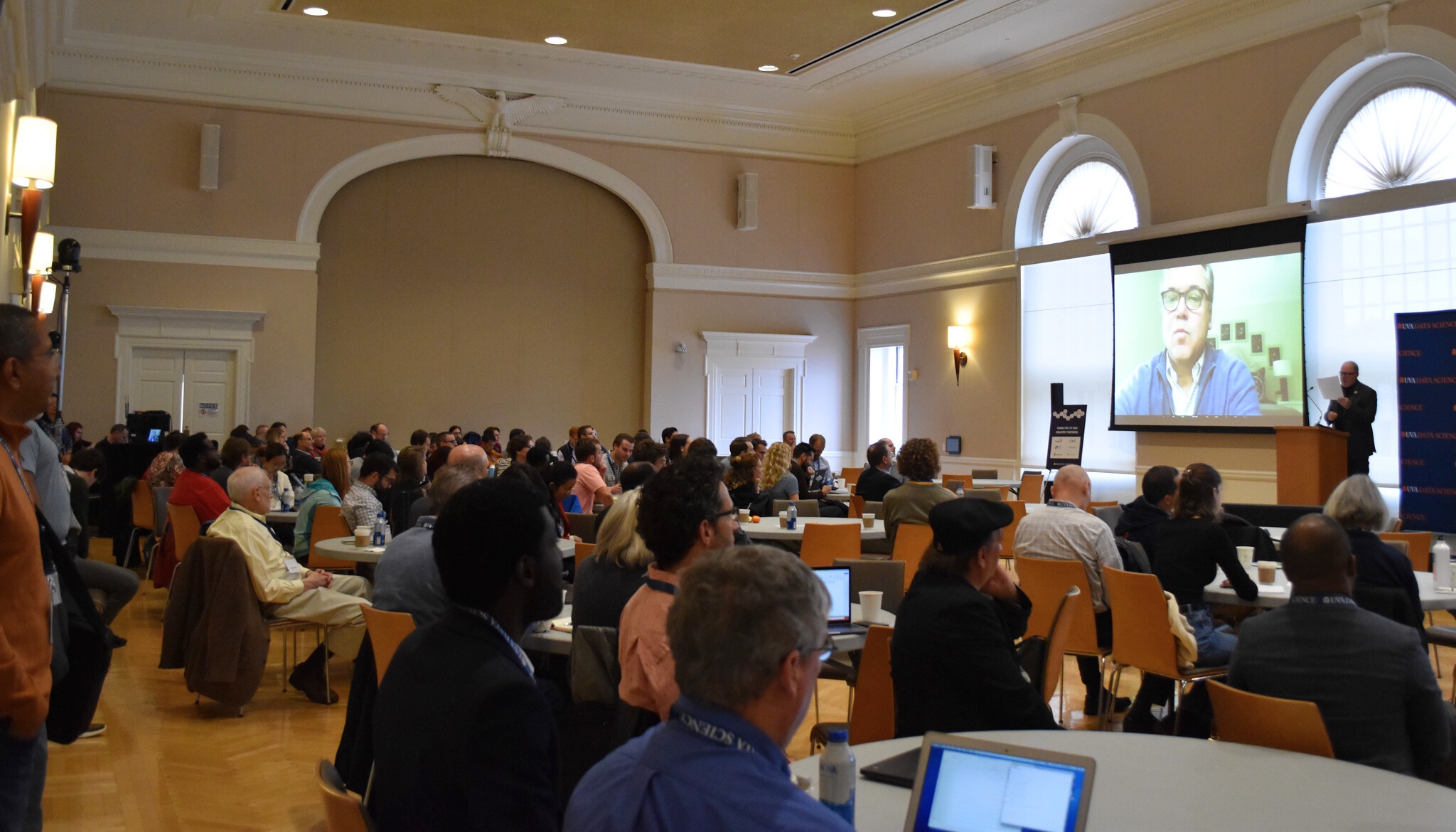 Audience seated in a conference room with high arched windows, watching a speaker on a large screen. Professional and focused atmosphere.