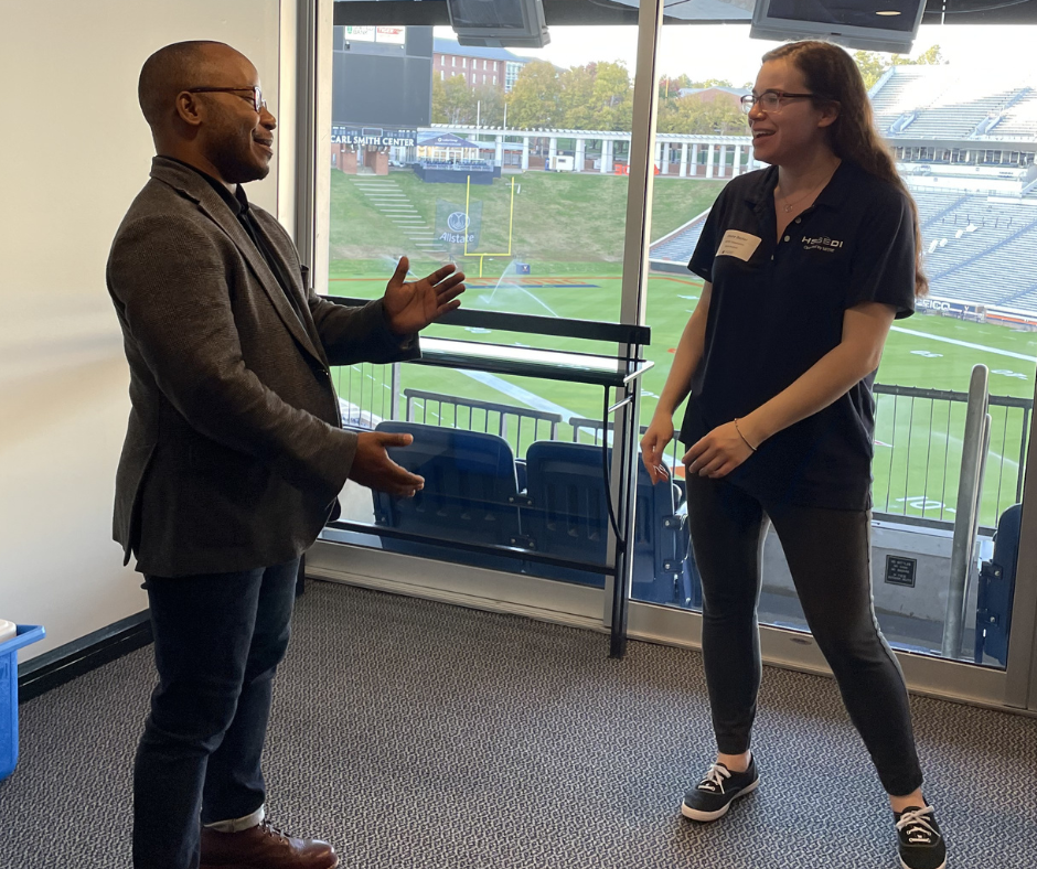 A man and a woman engage in a lively conversation in a room overlooking a stadium. Both appear animated and enthusiastic, creating a friendly atmosphere.