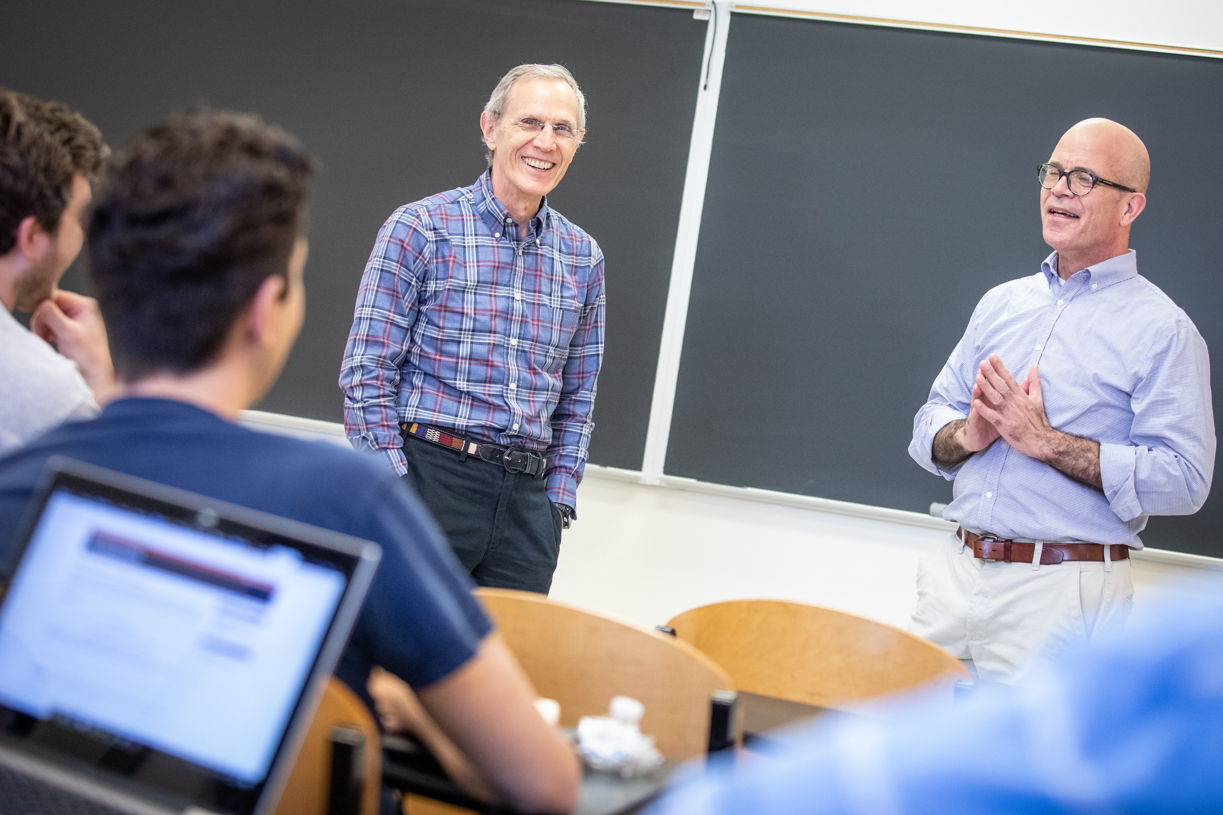 Two individuals stand in front of a classroom, discussing while students sit at desks, focused on the lecture.