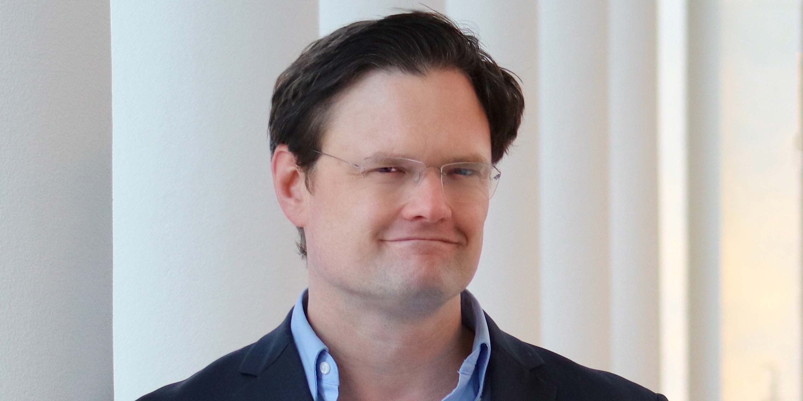 Man with short dark hair wearing glasses and a blue shirt under a black blazer, standing indoors with a neutral expression, in front of white columns.