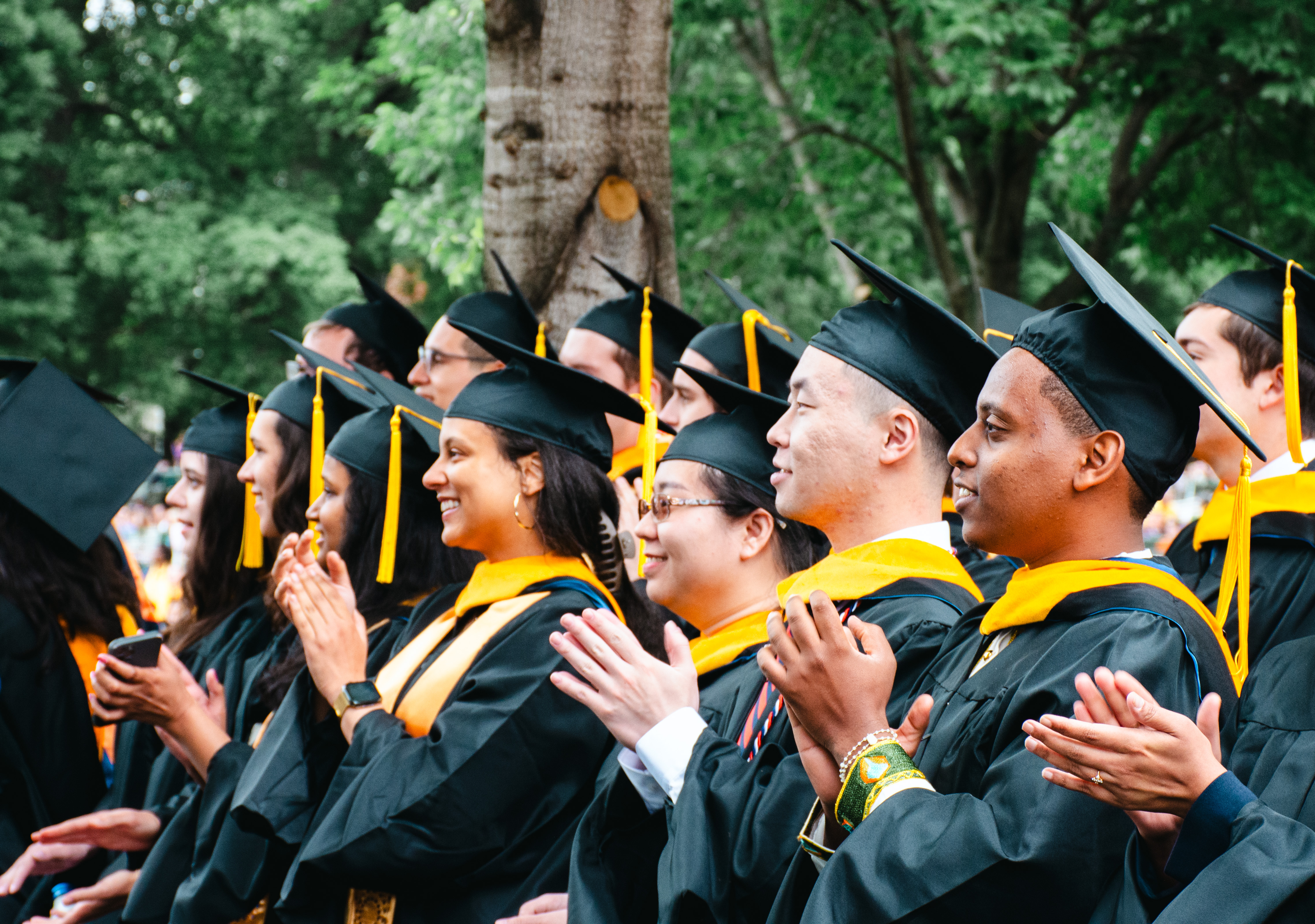 Graduates in caps and gowns applaud during a commencement ceremony, surrounded by greenery and a celebratory atmosphere.