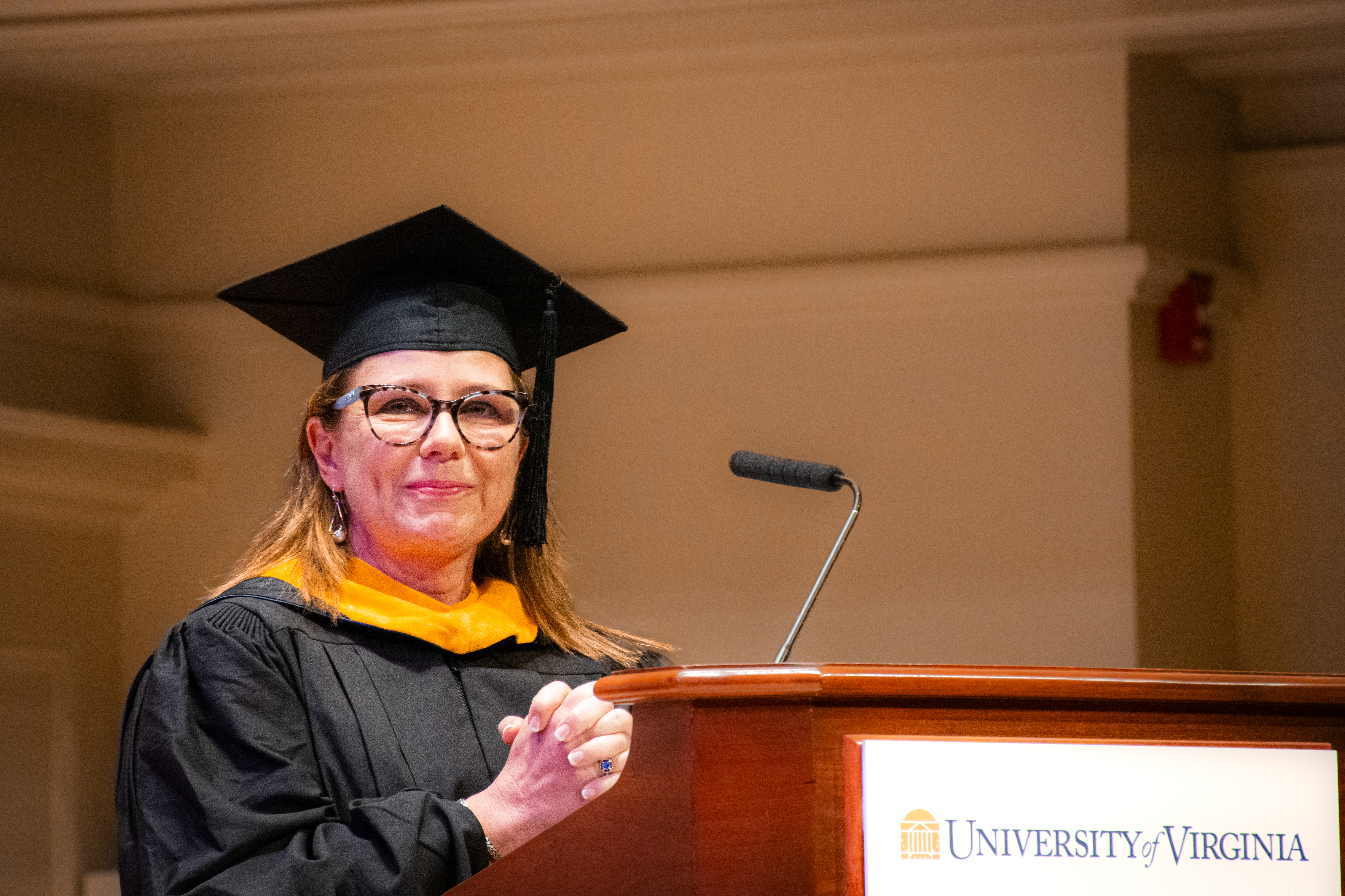 Woman in graduation attire delivers a speech at a podium with a University of Virginia sign. She looks confident and joyful, capturing the ceremony's celebratory tone.