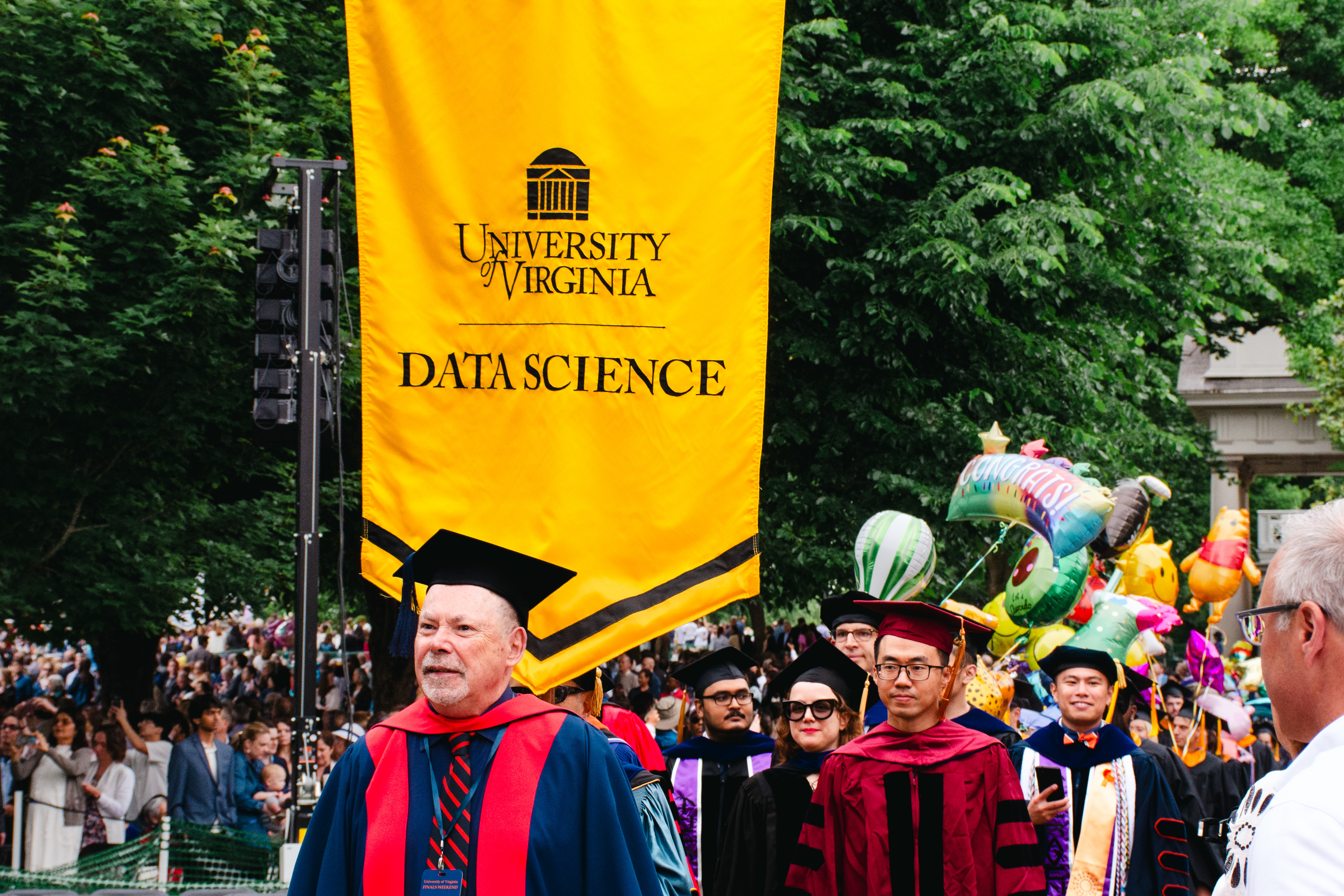 Graduates in colorful gowns and caps walk under a yellow "University of Virginia Data Science" banner on a festive, tree-lined campus path.