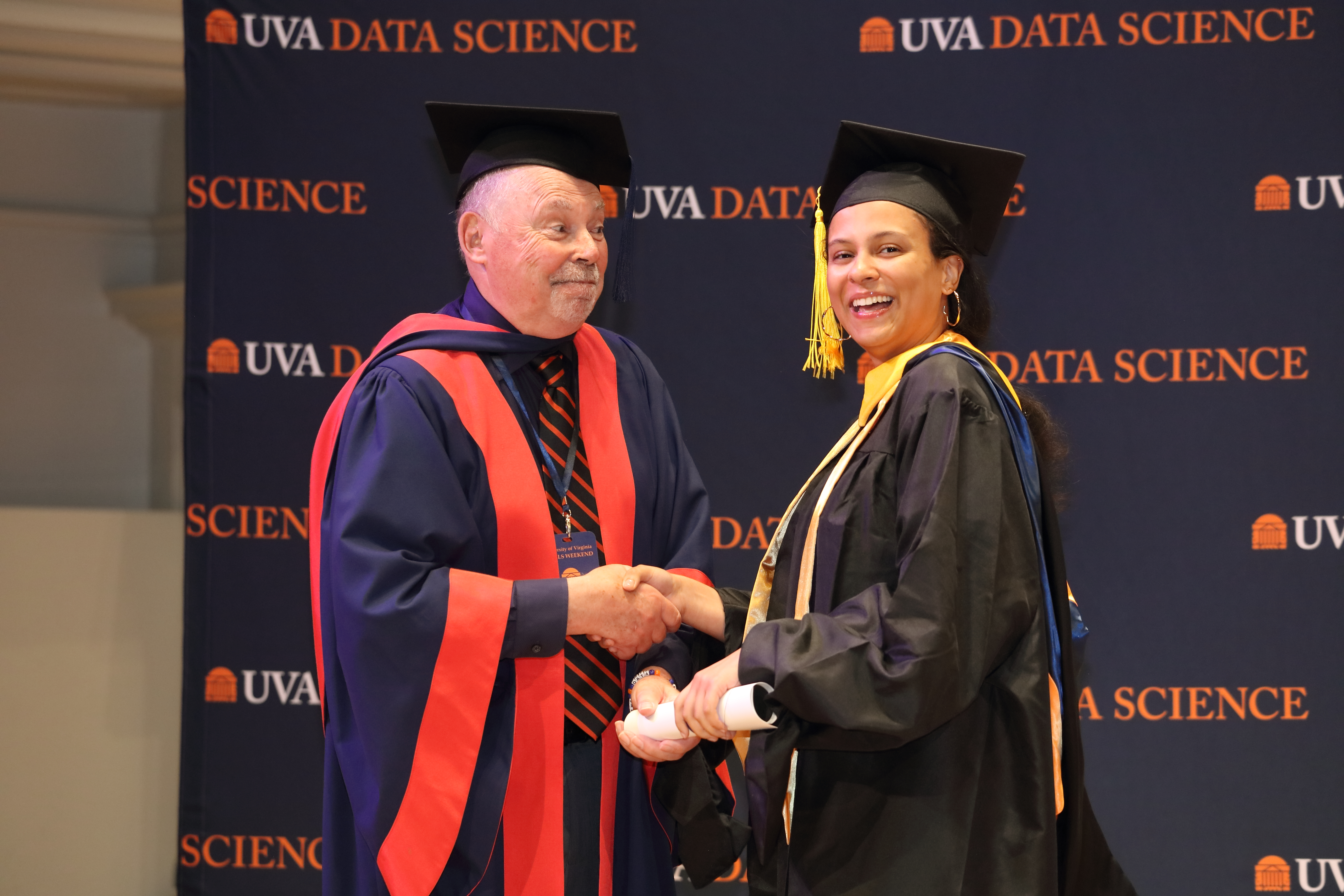 A graduate in a black cap and gown shakes hands with a faculty member in academic regalia. Both are smiling in front of a UVA Data Science backdrop.