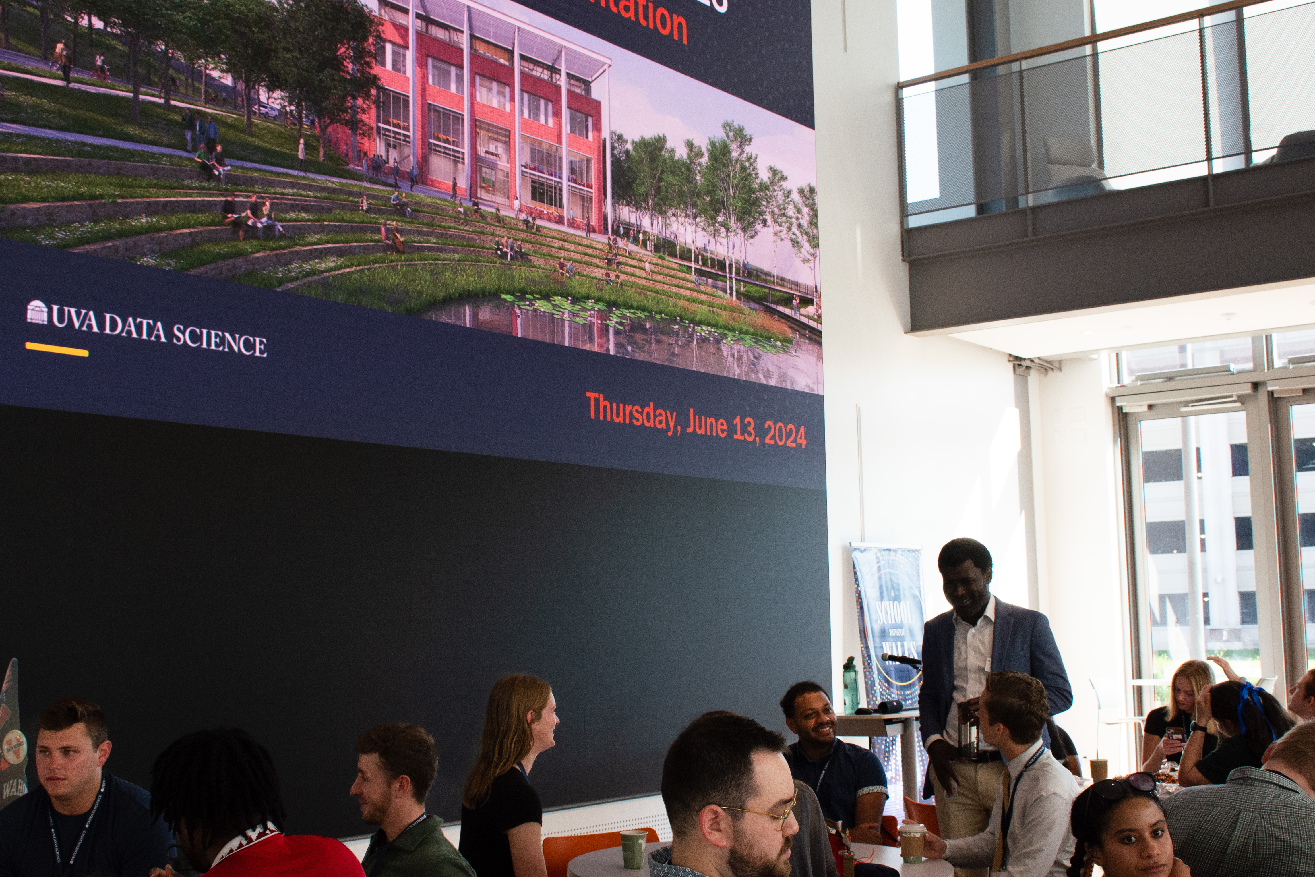 A group of people in a modern atrium gather beneath a large digital display showing "UVA Data Science" and "Thursday, June 13, 2024." 