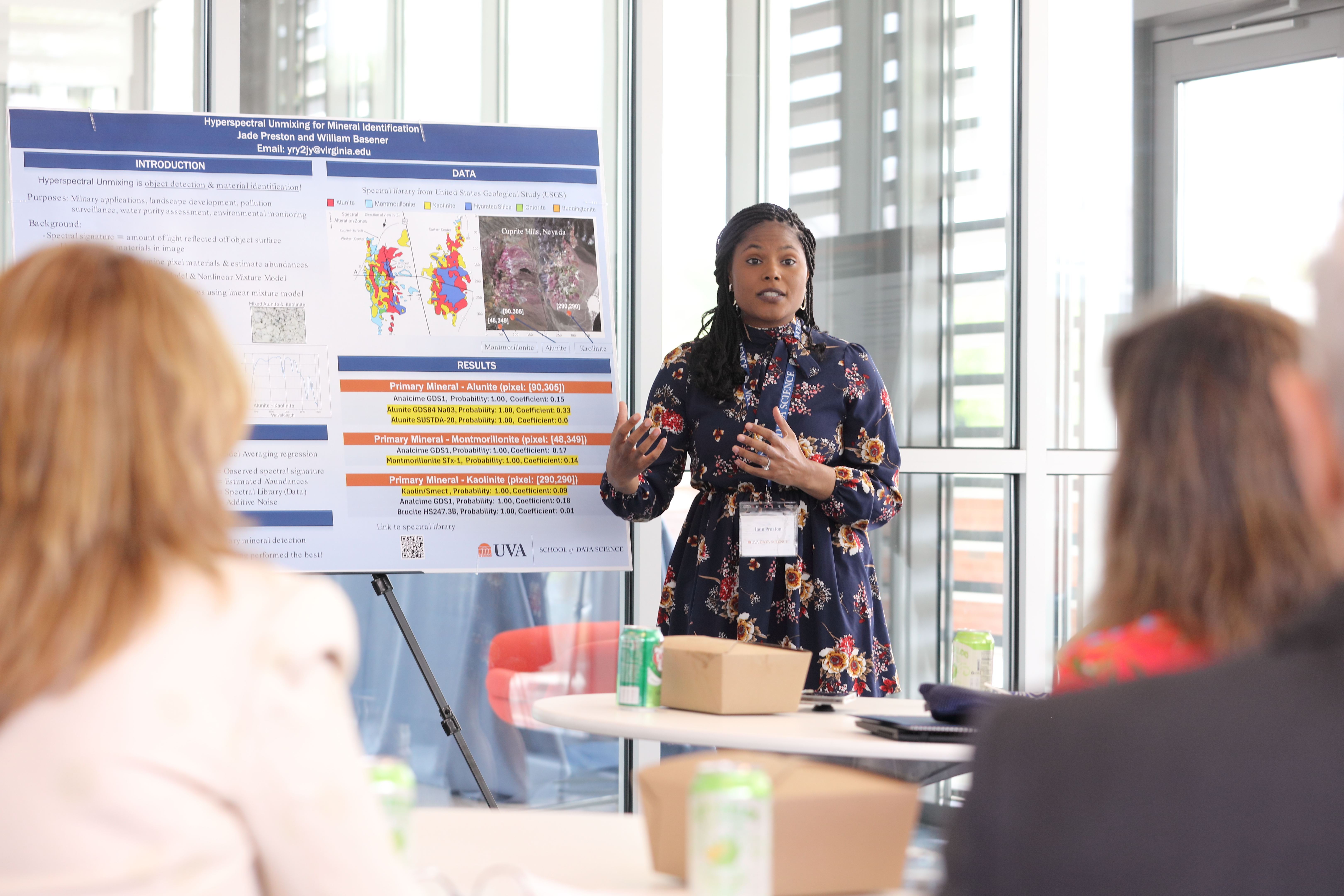 A woman presents research in front of a colorful poster at a conference. She gestures engagingly, while attendees listen attentively in a bright room.