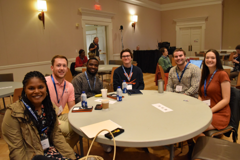 A group of six people is sitting around a table in a conference room, smiling at the camera. They have notebooks, water bottles, and name tags.