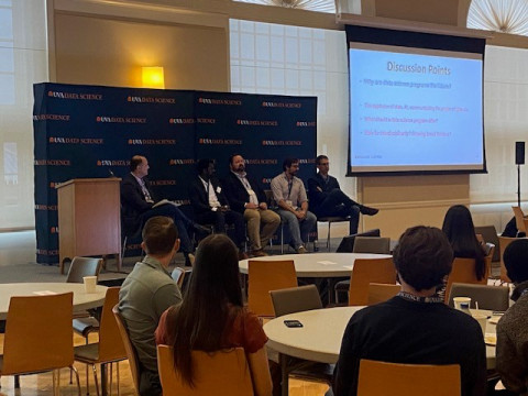 Panel of five speakers on a stage with "UVA Data Science" backdrop, discussing topics on a screen. Audience seated at round tables, attentive and engaged.