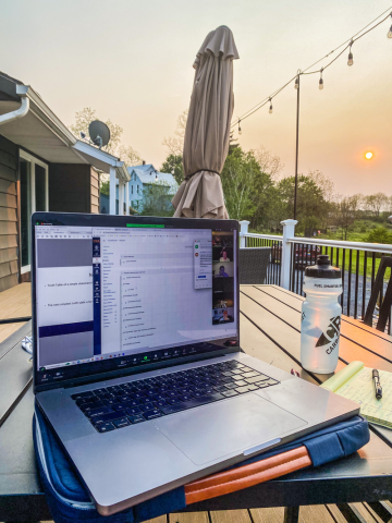 A laptop displaying a video call is on a wooden patio table beside a notebook and a water bottle. The background shows a sunset and garden view.