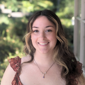 Young woman smiling warmly, standing in soft focus garden. Wearing a floral dress, necklace, and wavy hair with sunlit greenery in the background.