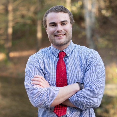 Photo of Bryan Christ wearing a button up blue shirt and red tie standing with arms crossed and smiling in front of a blurred nature background