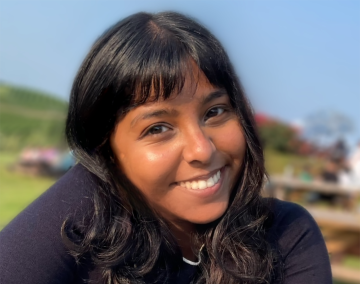 A person with long dark hair and a friendly smile is outdoors on a sunny day. The background is blurred, showing greenery and people in the distance.