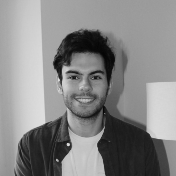 A person stands in front of a gray wall, wearing a dark jacket over a white shirt, with a lamp visible to the right.