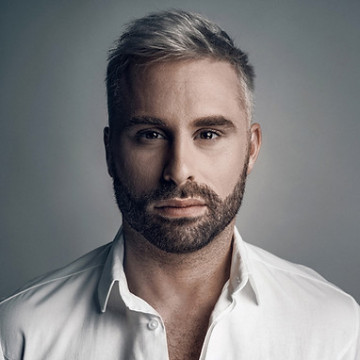 A man with short, light hair and a trimmed beard gazes intently at the camera. He wears a white collared shirt, set against a soft, neutral background.