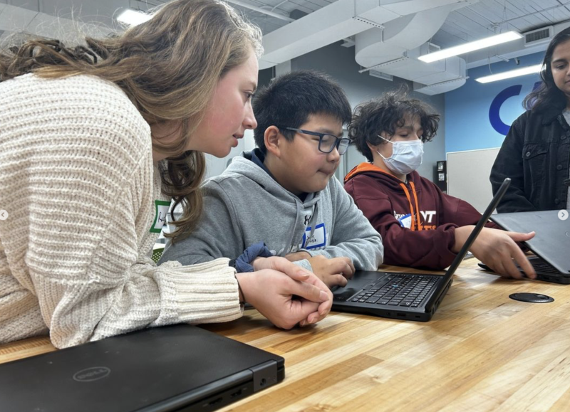 Group of students working together at a wooden table with laptops. Two focus intently on a screen, one wears a mask.