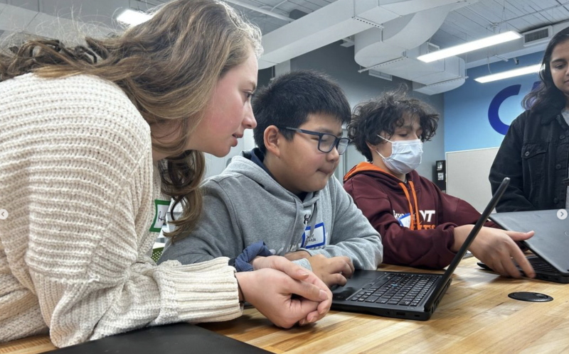 A group of four students collaborates around a laptop at a wooden table. One wears a mask, engaged and focused in casual, well-lit classroom.