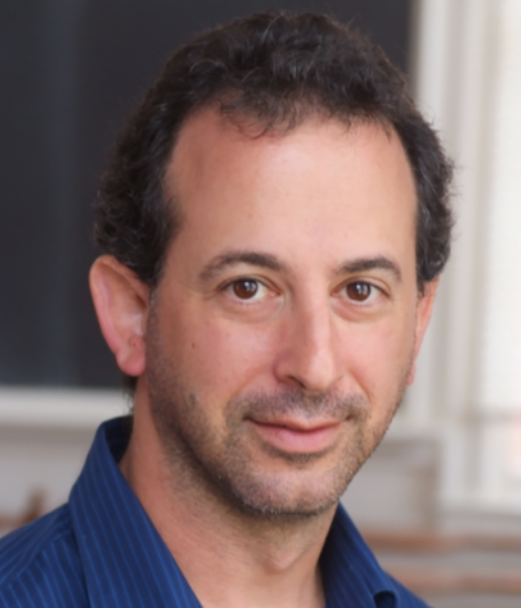 A middle-aged man with short, curly dark hair and a light beard, wearing a blue shirt. He looks directly at the camera with a slight smile, indoors.