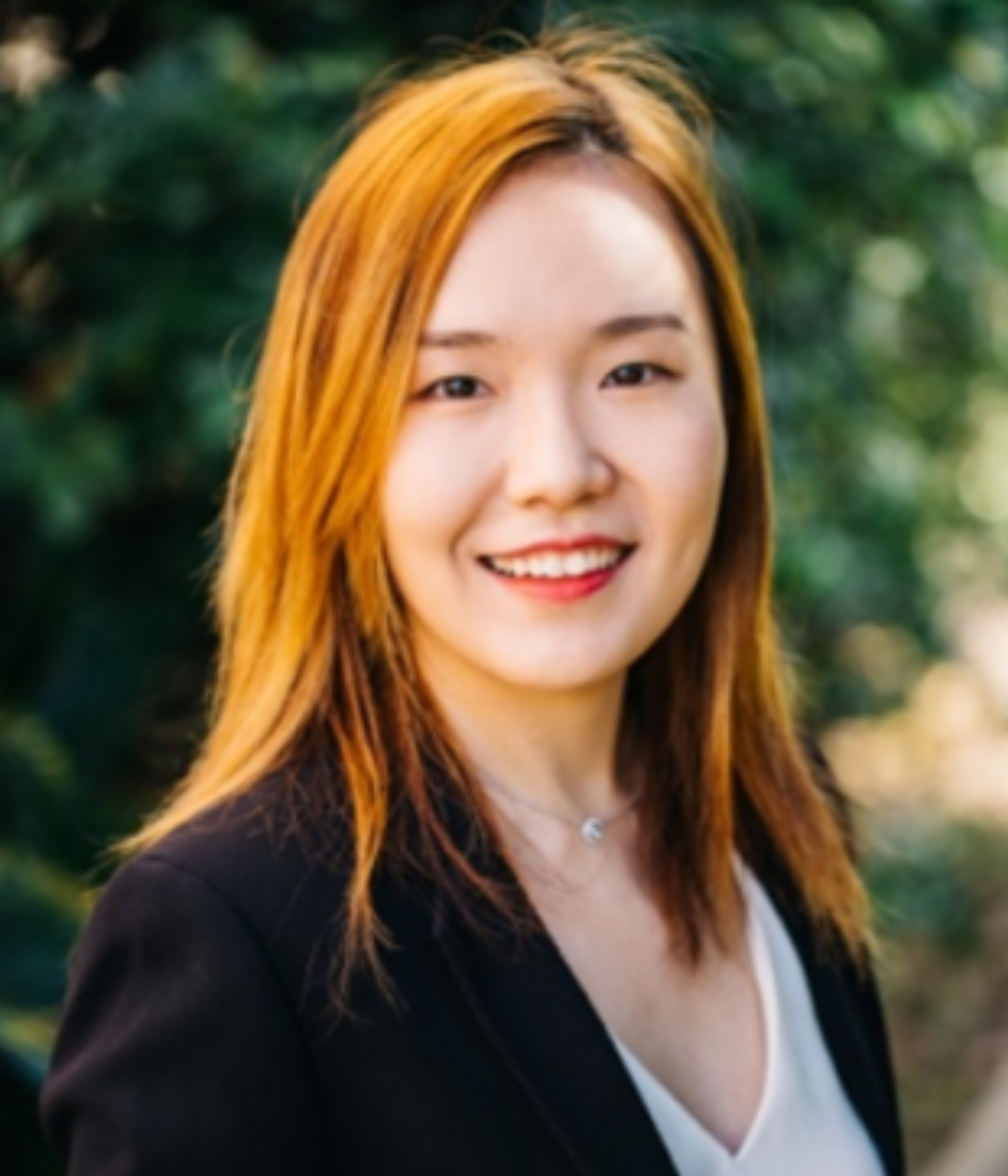 A woman wearing a white blouse and black blazer is smiling outdoors with green trees behind her.