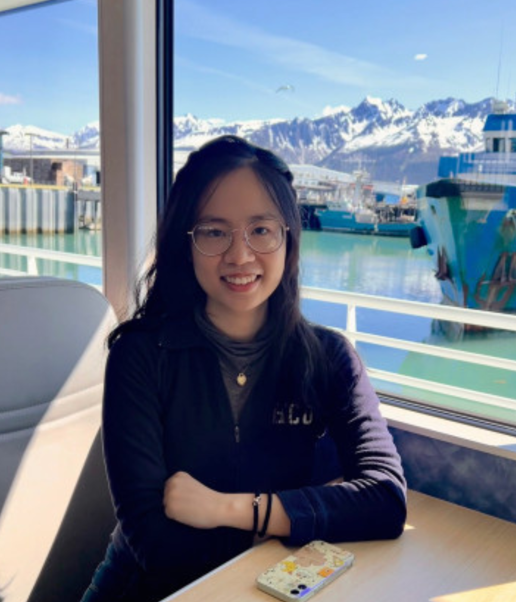 A woman with glasses smiles, seated indoors by a window. Outside, a harbor with docked boats and snow-capped mountains under a blue sky is visible.