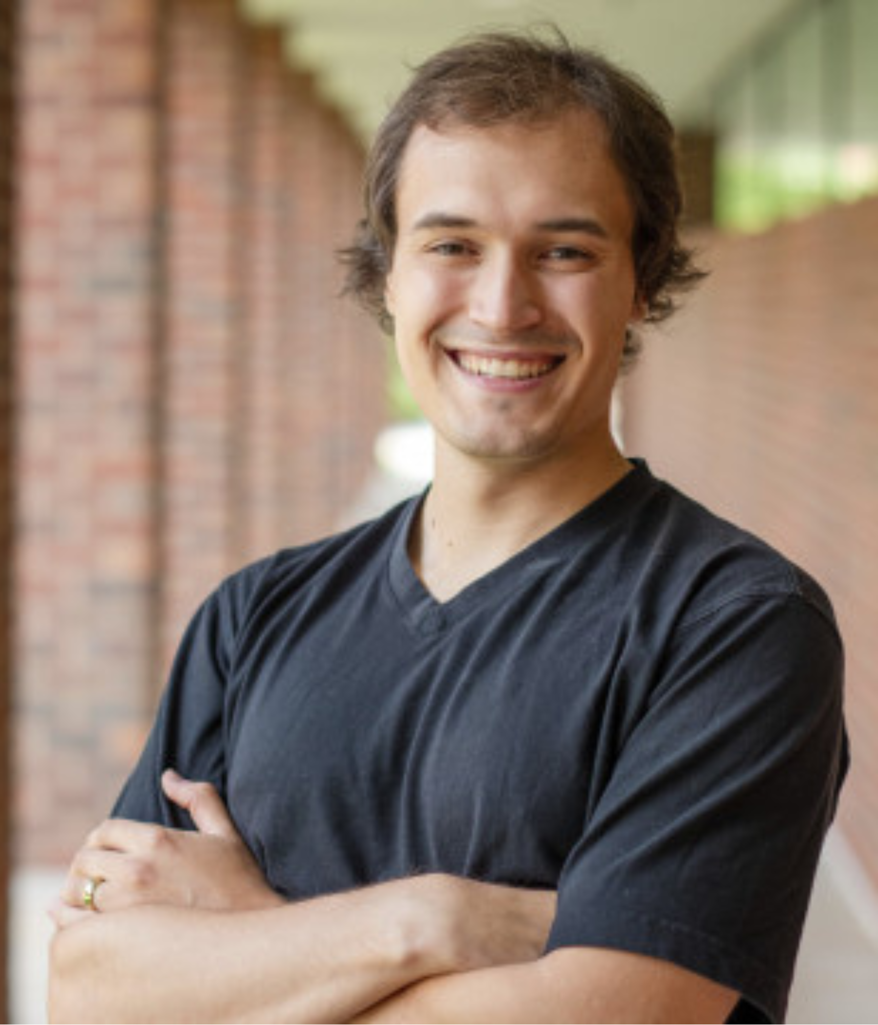 A smiling person with crossed arms wearing a black T-shirt stands in an outdoor hallway with brick walls. 