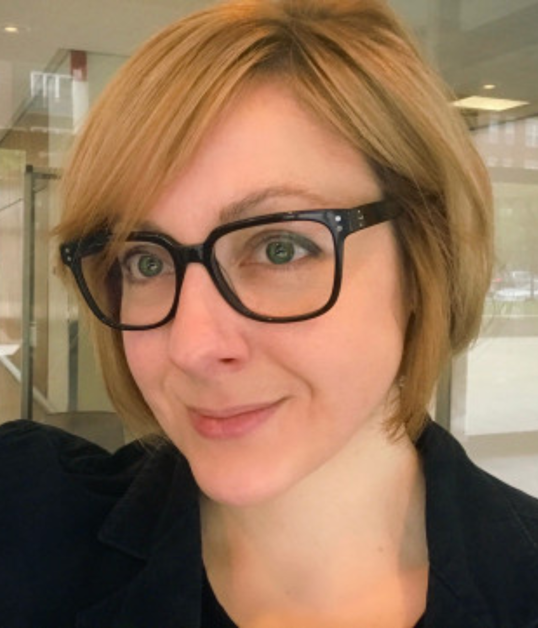 A person with short, blonde hair and wearing black glasses smiles softly indoors. 