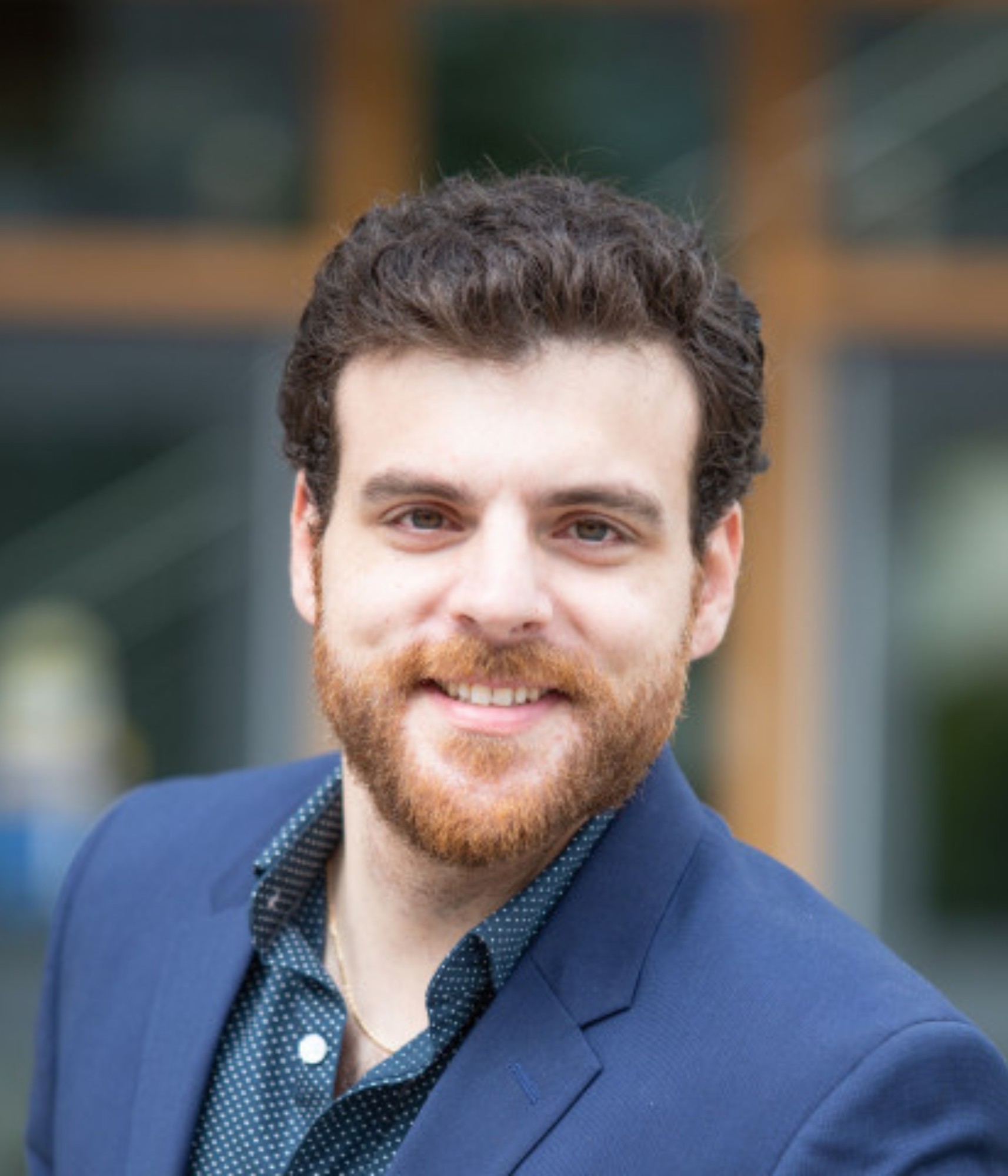 A smiling man with curly hair and a beard wears a blue blazer over a dotted shirt.   A man dressed in a blue blazer and polka dot shirt stands outdoors, with a blurred background of a modern building.