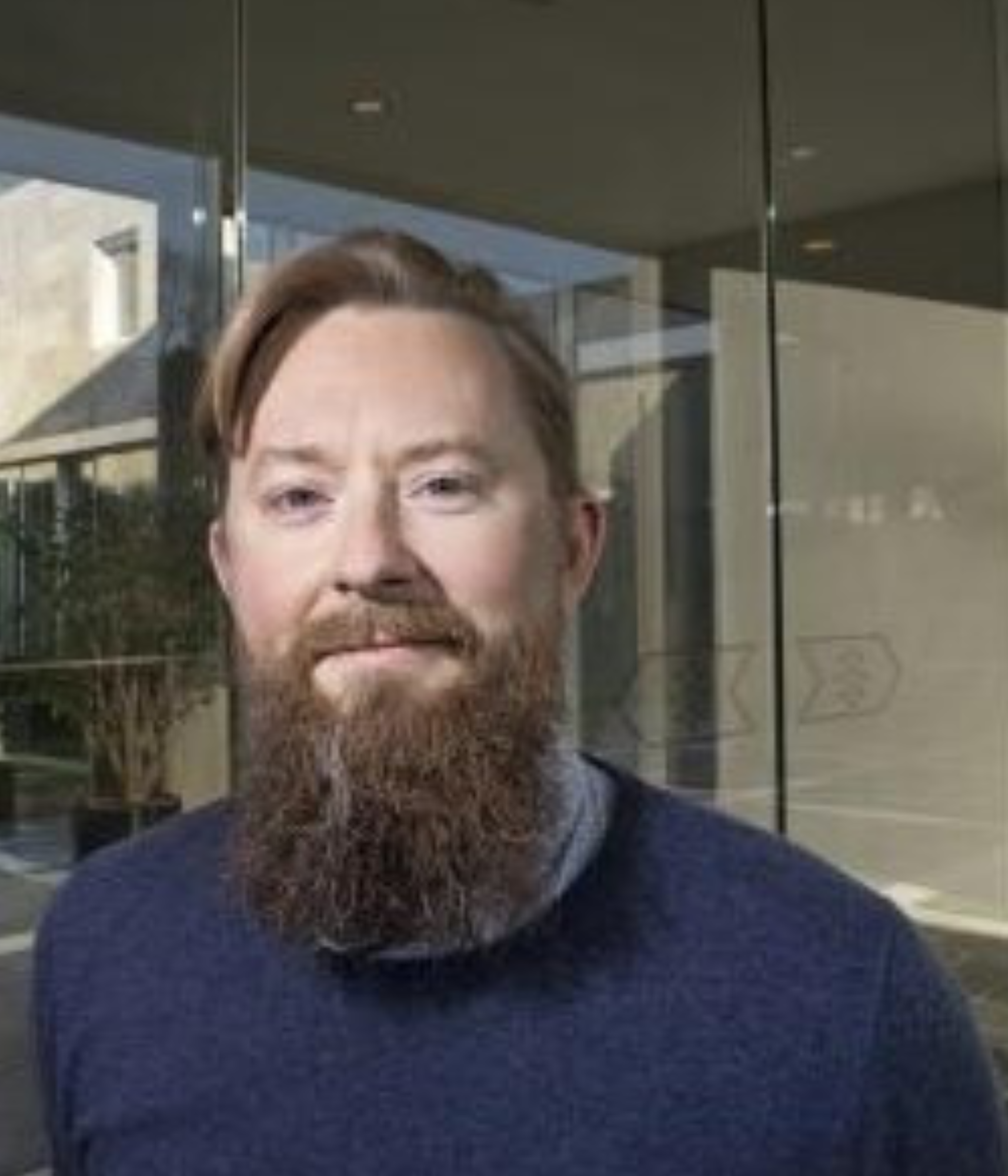 A man with a beard and blue sweater stands smiling in front of a glass building. 