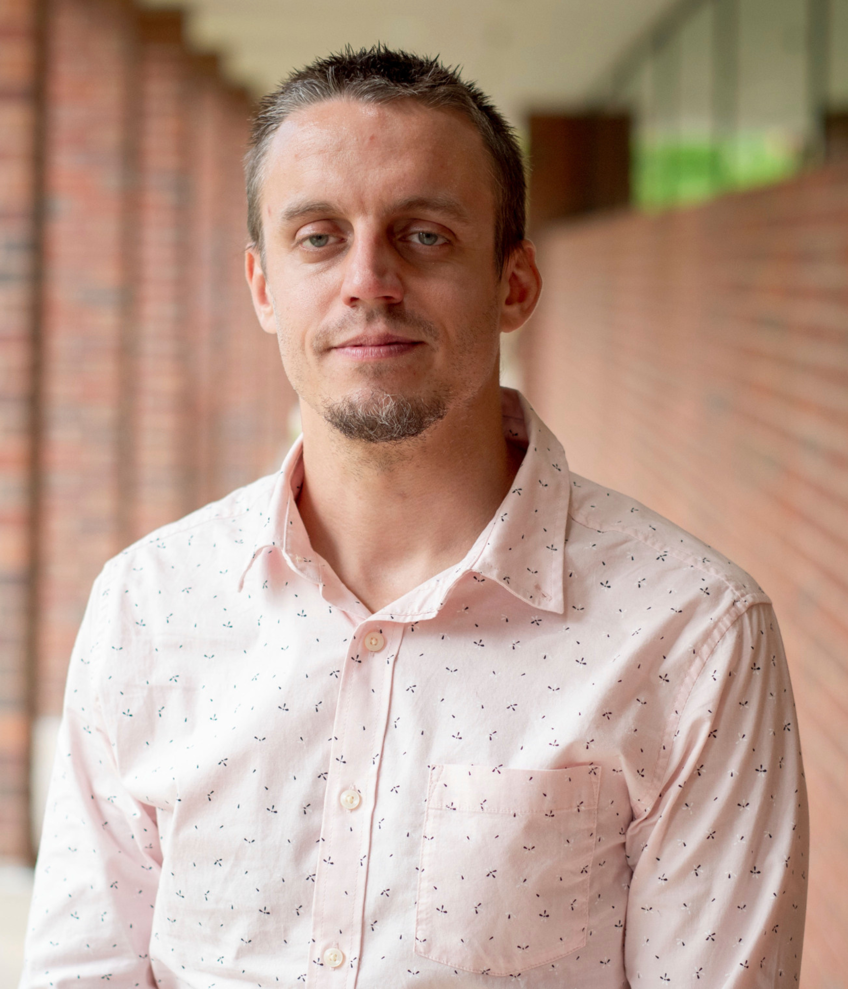 A man with short hair and a goatee wears a light pink, patterned shirt, standing against a blurred brick wall background with a calm expression.