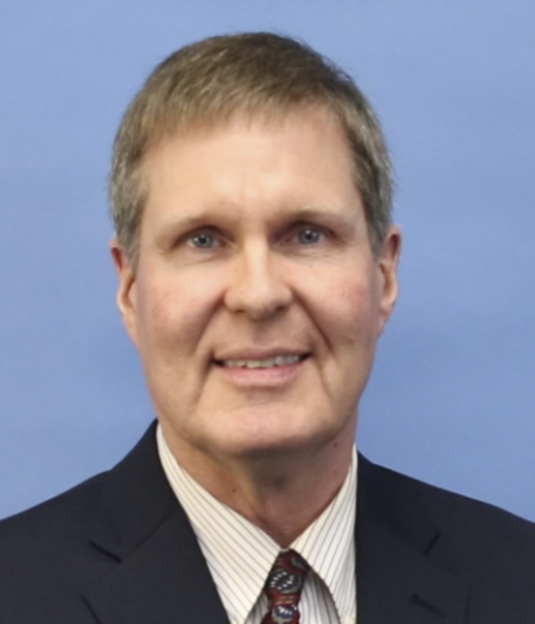 A middle-aged man with short light brown hair, wearing a dark suit, striped shirt, and patterned tie, smiles gently against a plain blue background.