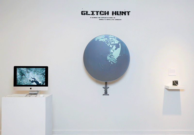Exhibit titled "Glitch Hunt" shows a wall installation with a digital globe and a satellite image on a computer. Minimalist, tech-focused art display.