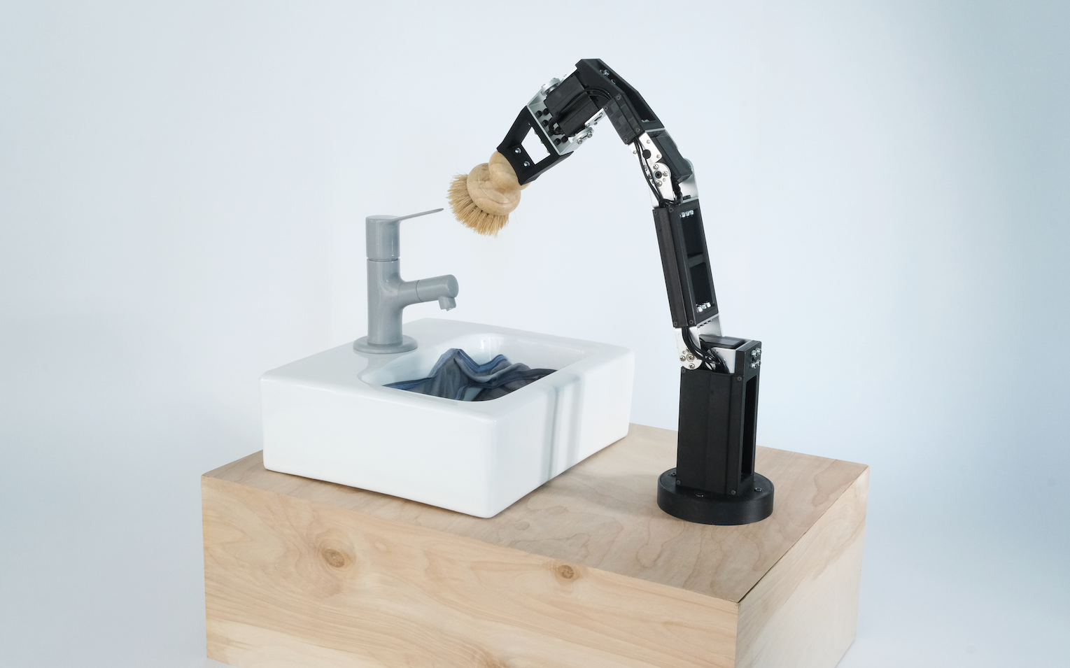 A black robotic arm with a wooden brush cleans a cloth in a white sink with a gray faucet, set on a wooden surface, in a minimalist setting.