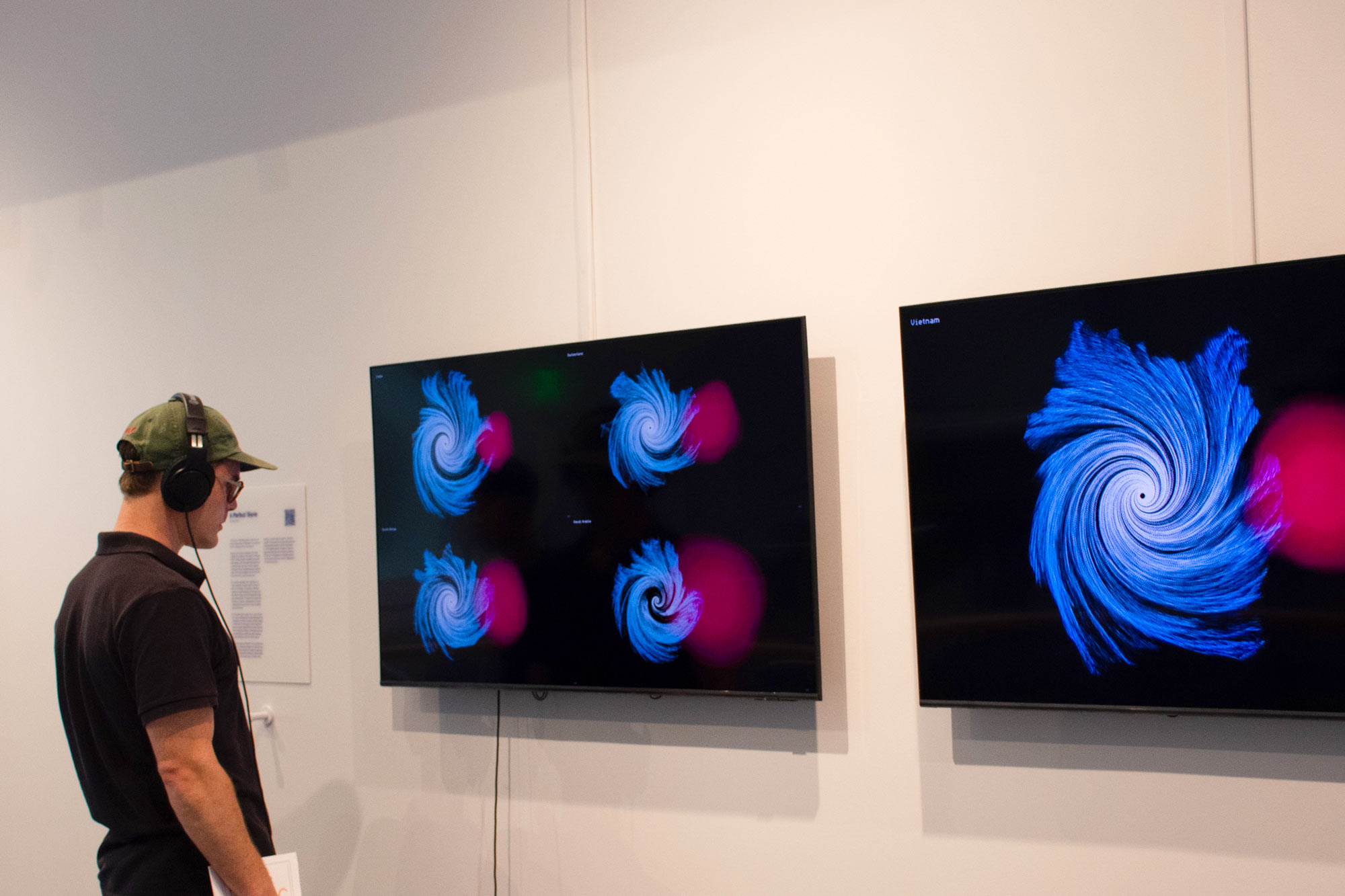 A man wearing headphones observes two wall-mounted screens displaying vibrant abstract art. The art features swirling blue patterns with bright pink accents.