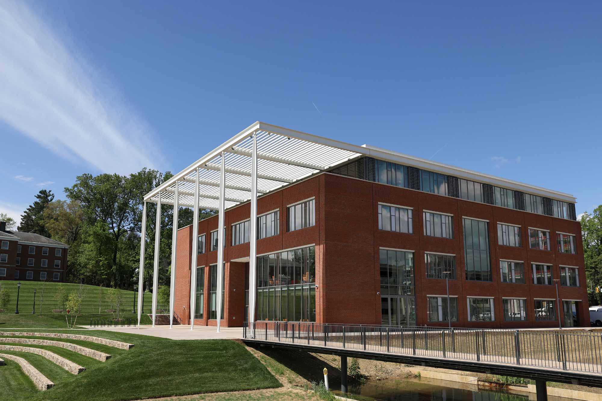 A modern, red-brick building with large windows and white columns under a clear blue sky, surrounded by green lawns and trees.