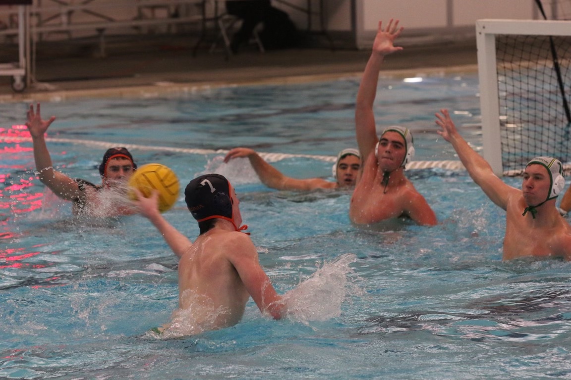A water polo player in a dark cap prepares to throw a yellow ball towards the goal, while four defenders with raised arms attempt to block.