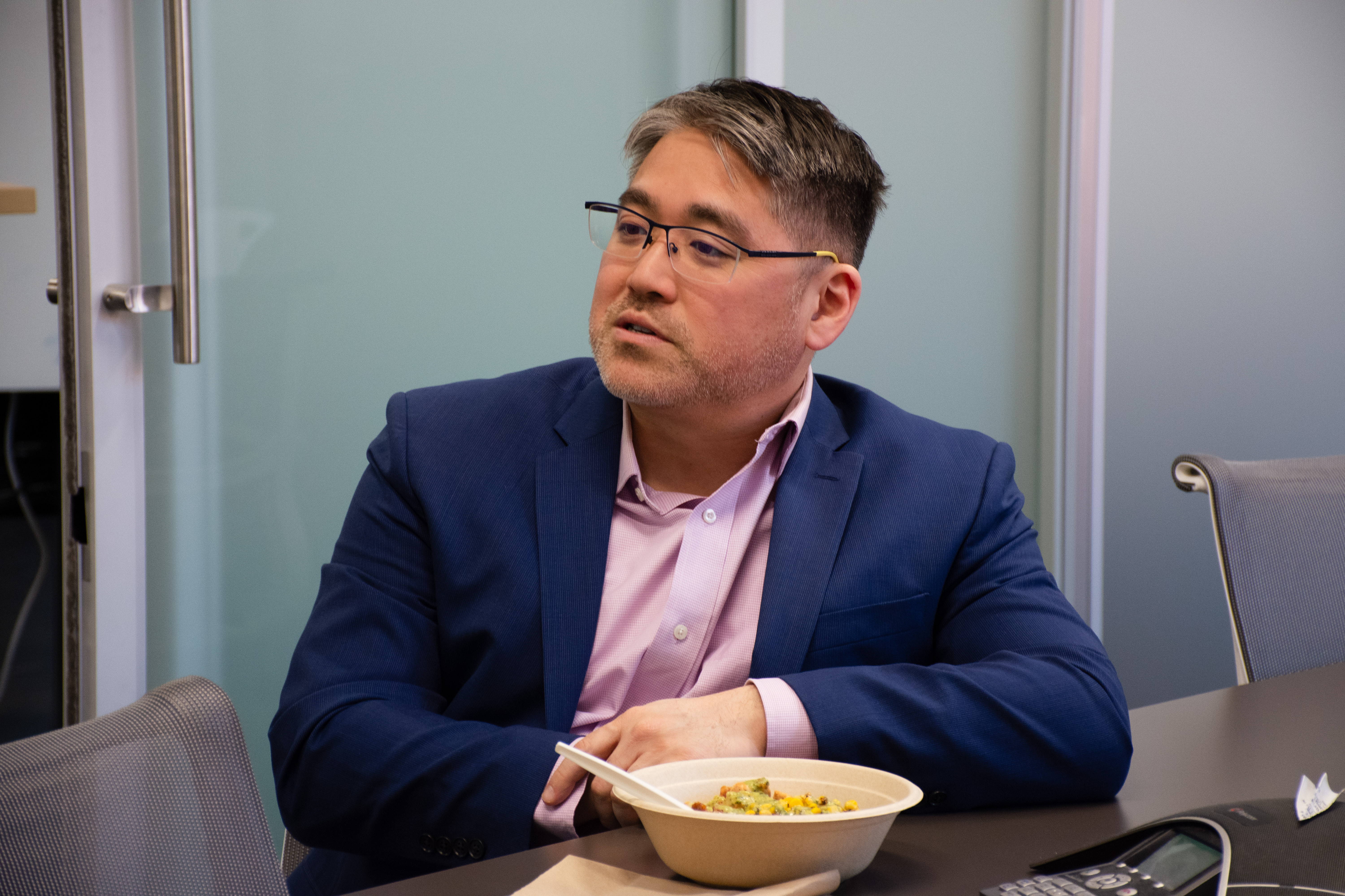 Man in a blue suit, pink shirt, and glasses sits at a conference table with a bowl of food, conveying focus and engagement in a discussion.