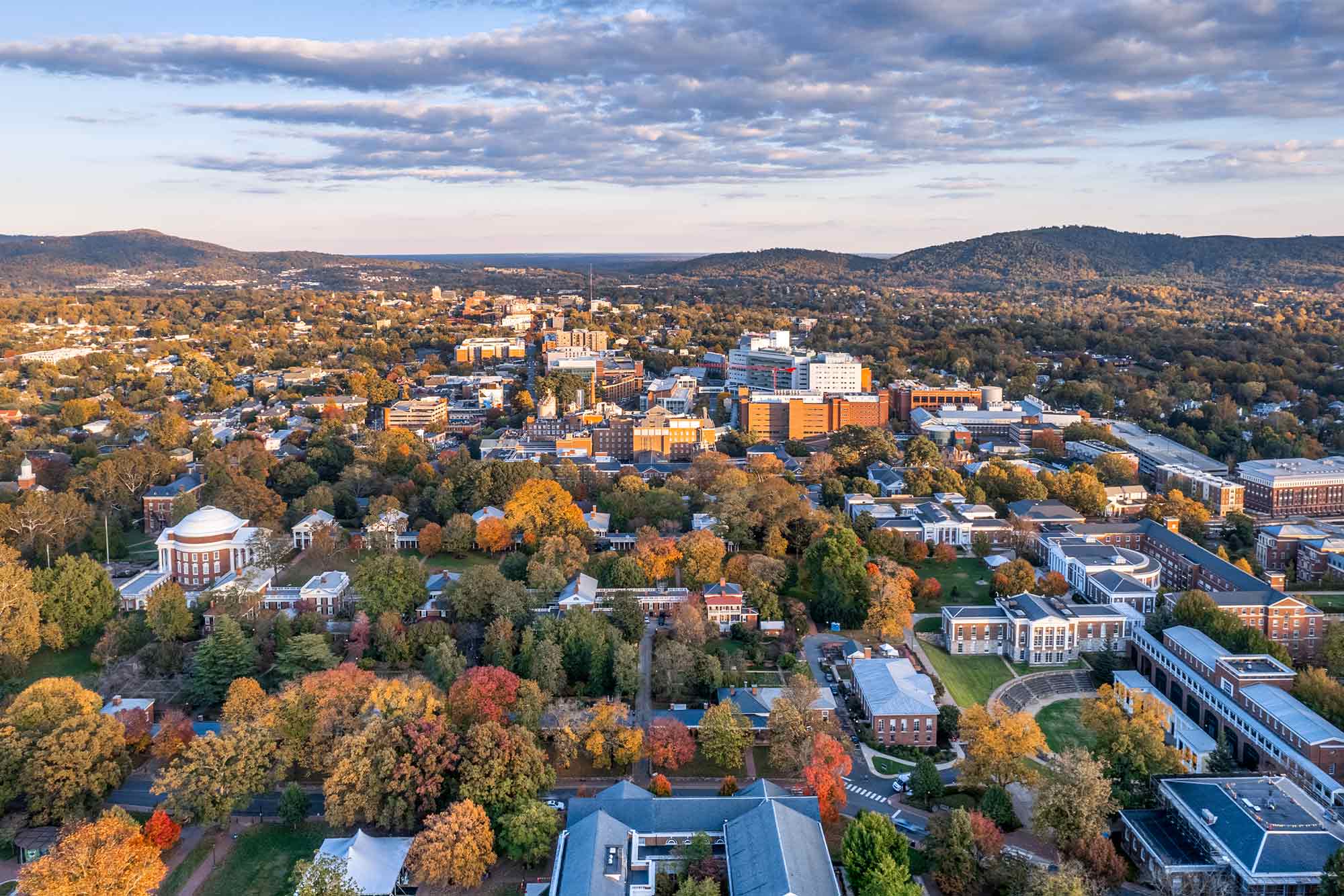 Aerial view of a sprawling university campus in autumn, surrounded by colorful trees and hills in the background under a partly cloudy sky.