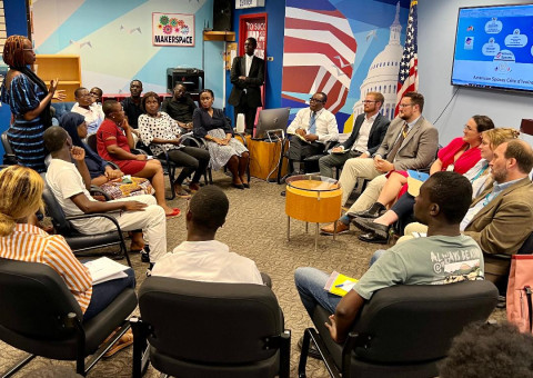 A diverse group of around twenty people in a room sit in a circle, attentively listening to a speaker. The room has colorful wall art, a U.S. flag, and a presentation screen displaying slides.