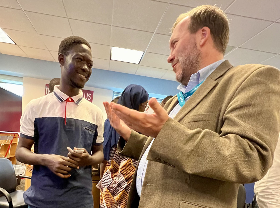 A smiling man in a polo shirt listens to a bearded man in a jacket gesturing animatedly. Others converse in the background.