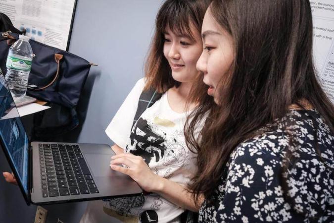 Two women are smiling and looking at a laptop screen. They appear engaged and focused. A bottle and bag are nearby on a table, suggesting a casual setting.