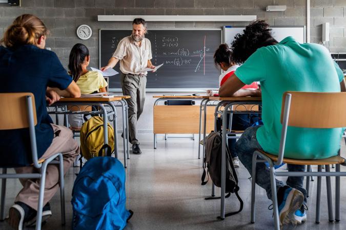 Classroom scene with a male teacher holding papers, explaining math on a chalkboard. Students sit attentively at desks; a clock displays midday.