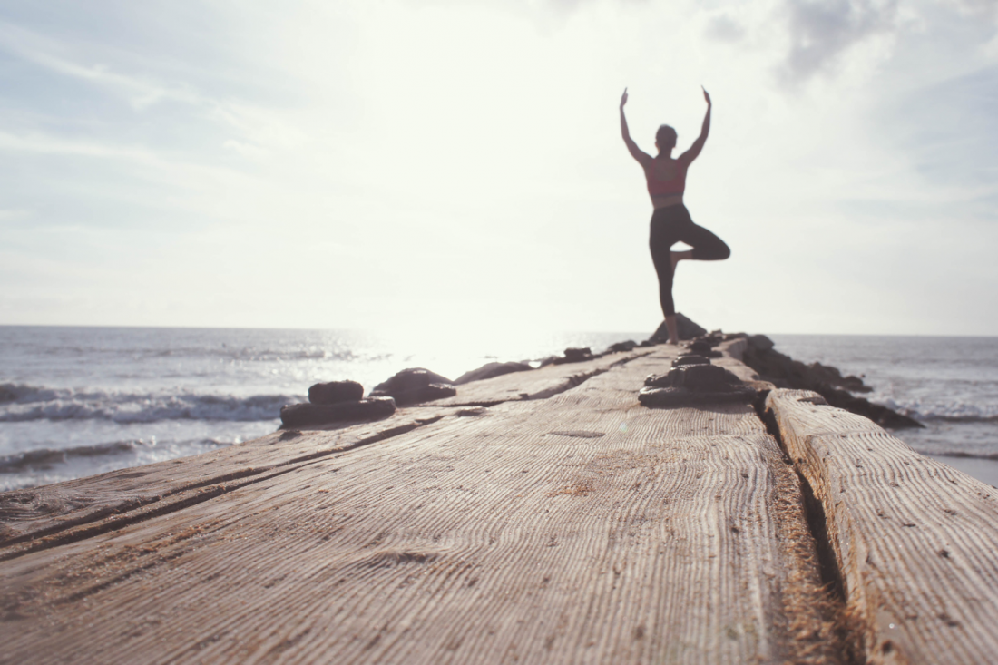 Silhouette of a person doing yoga in tree pose on a wooden pier, facing the ocean. 