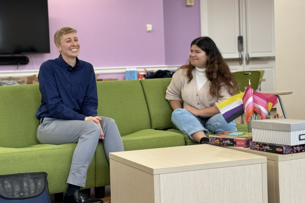 Two people are sitting on a green couch in a room with purple walls, smiling and talking. Nearby are pride flags and board games on a table. 