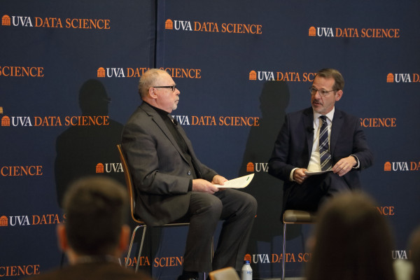 Two men in suits are seated on a stage with "UVA Data Science" banners in the background. They are conversing seriously, holding papers. Audience blurs in foreground.