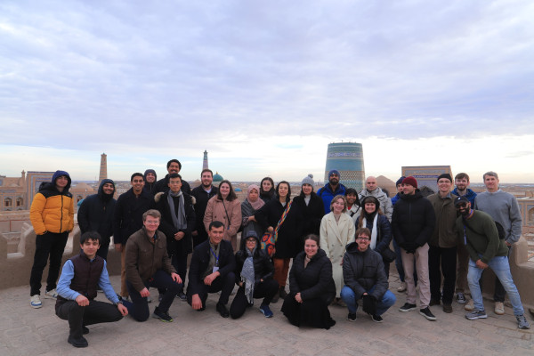 A large group poses together on a scenic overlook, with historic towers and a vast landscape in the background under a cloudy sky.