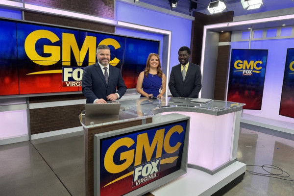 Two news anchors stand smiling behind a clear desk with a guest behind them; text reads "GMC FOX Virginia" on a screen in the background. They are in a modern TV studio.