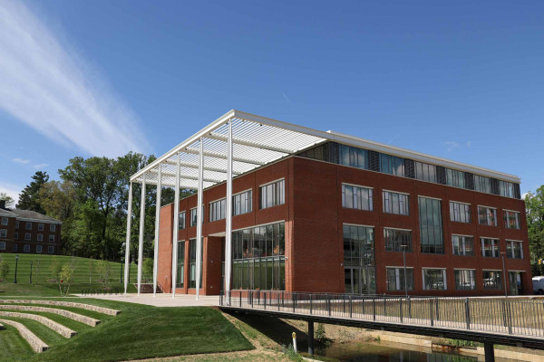 A modern, red-brick building with large windows and white columns under a clear blue sky, surrounded by green lawns and trees.