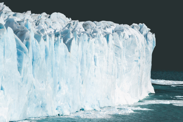 A massive icy glacier with a rugged surface, deep blue crevices, and towering edge against a dark sky, meeting the calm teal ocean below. 