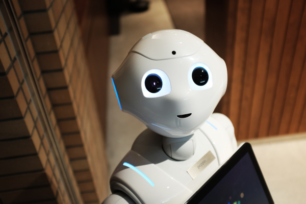 A humanoid robot with a smooth white surface, expressive eyes, and a small screen, standing indoors near brick and wood walls.