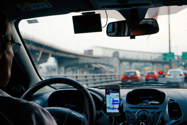 A person driving a car in heavy traffic, holding the steering wheel. A GPS navigation app is displayed on a phone mounted on the dashboard.